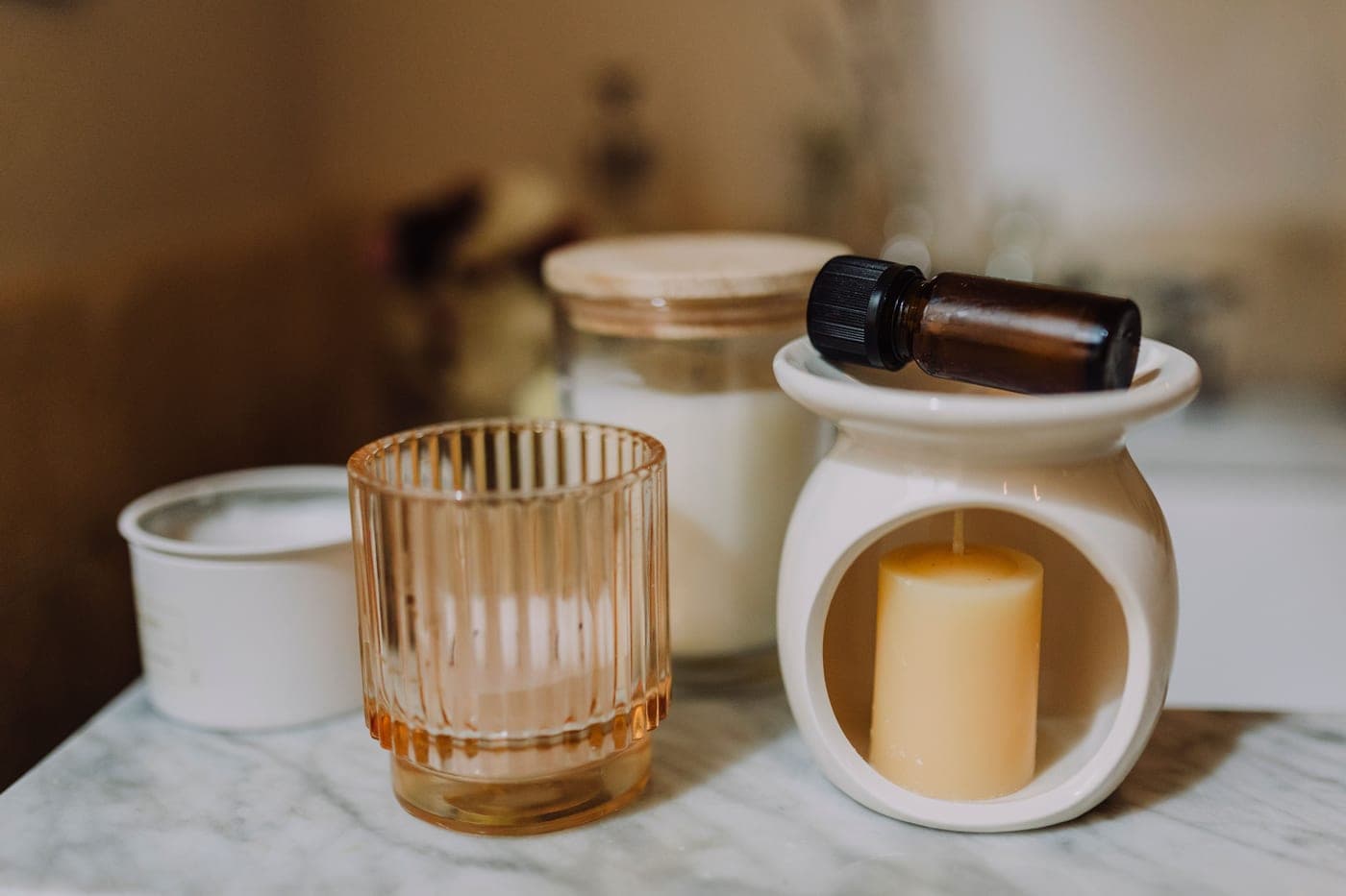 A white ceramic wax burner with an unlit block of wax in it, beside unlit candles and a bottle of fragrance oil on a marble surface